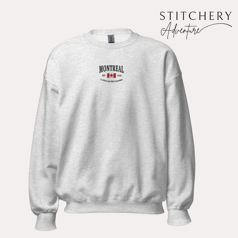 May include: A light gray sweatshirt with the word "MONTREAL" in black text above a red maple leaf and the text "EST. 1642". The sweatshirt has a crew neck and long sleeves. The brand name "STITCHERY Adventure" is in the upper right corner.