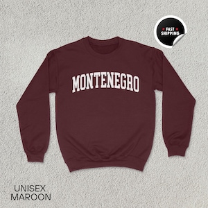 May include: Maroon unisex sweatshirt with the word "MONTENEGRO" in white, arched lettering across the chest. The sweatshirt has long sleeves and a crew neck. The words "UNISEX MAROON" are printed at the bottom. A "FAST SHIPPING" sticker is in the upper right corner.