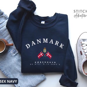 May include: A navy blue crewneck sweatshirt featuring the white text DANMARK in an arched font, a small crown emblem, two crossed Danish flags, and the text KØBENHAVN 55.6761 N, 12.5683 E. The sweatshirt is displayed with light blue jeans and grey trainers.
