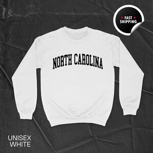 May include: A white unisex sweatshirt with the text "NORTH CAROLINA" in black, arched lettering. The sweatshirt has long sleeves and a crew neck. The words "UNISEX WHITE" are printed at the bottom. A sticker that says "FAST SHIPPING" is in the upper right corner.