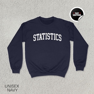 May include: A navy blue sweatshirt featuring the word "STATISTICS" in large, white, block letters. The garment has a crew neck and long sleeves. The words "UNISEX NAVY" are printed at the bottom. A "Fast Shipping" sticker is in the upper right corner.