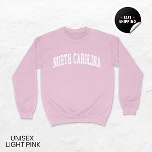 May include: A light pink unisex sweatshirt with the text "NORTH CAROLINA" in white, arched across the chest. The sweatshirt has long sleeves and a crew neck. The words "UNISEX LIGHT PINK" are at the bottom.