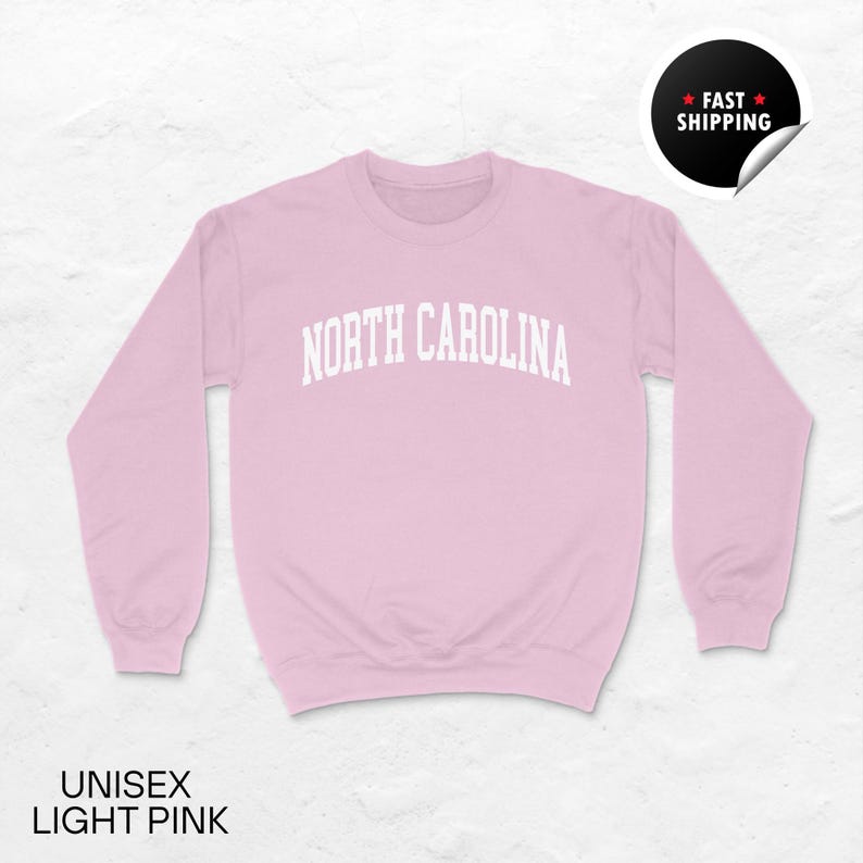 May include: A light pink unisex sweatshirt with the text "NORTH CAROLINA" in white, arched across the chest. The sweatshirt has long sleeves and a crew neck. The words "UNISEX LIGHT PINK" are at the bottom.