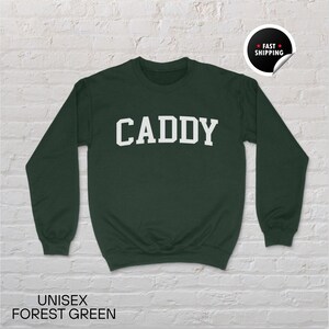 May include: Forest green unisex sweatshirt with the word "CADDY" in white block letters. The sweatshirt has a crew neck and long sleeves. The text "UNISEX FOREST GREEN" is printed at the bottom of the image.