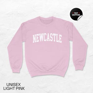 Puede incluir: Sudadera unisex rosa claro con la palabra "NEWCASTLE" en letras blancas en el pecho. La sudadera tiene mangas largas y cuello redondo. El texto "UNISEX LIGHT PINK" est&aacute; en la parte inferior de la imagen. Una pegatina de "FAST SHIPPING" est&aacute; en la esquina superior derecha.