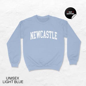 Puede incluir: Sudadera unisex azul claro con la palabra "NEWCASTLE" en letras blancas. La sudadera tiene mangas largas y cuello redondo. El texto "UNISEX LIGHT BLUE" est&aacute; en la parte inferior de la imagen. Una pegatina que dice "FAST SHIPPING" est&aacute; en la esquina superior derecha.