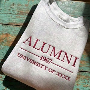 Può includere: Felpa grigio melange con ricamo bordeaux. La parola "ALUMNI" è sopra "1967" e "UNIVERSITY OF XXXX". Un rettangolo bordeaux nell'angolo in alto a sinistra recita "CUSTOM EMBROIDERED SHIRT". La felpa è piegata su una superficie di legno.