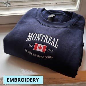 May include: A navy blue sweatshirt with white embroidered text reading "MONTREAL" above a Canadian flag and the text "EST 1642". Below is the text "LA VILLE AUX CENT CLOCHERS". The word "EMBROIDERY" is in a light blue box.