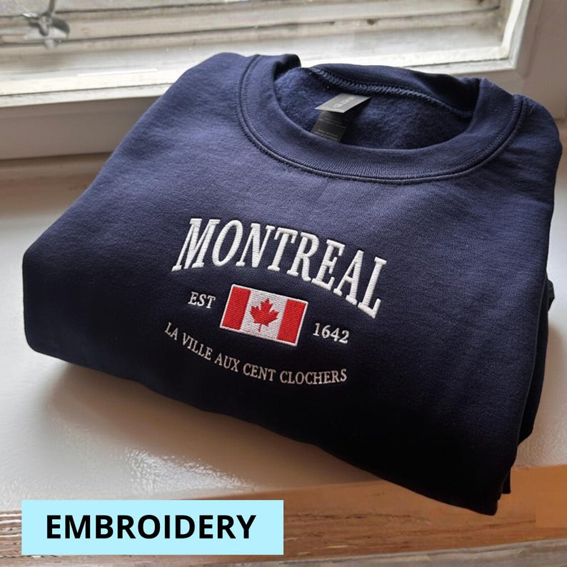 May include: A navy blue sweatshirt with white embroidered text reading "MONTREAL" above a Canadian flag and the text "EST 1642". Below is the text "LA VILLE AUX CENT CLOCHERS". The word "EMBROIDERY" is in a light blue box.