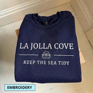 May include: Navy blue sweatshirt with white embroidered text that reads "LA JOLLA COVE" above a seal illustration and the text "KEEP THE SEA TIDY". The word "EMBROIDERY" is in a light blue box at the bottom.