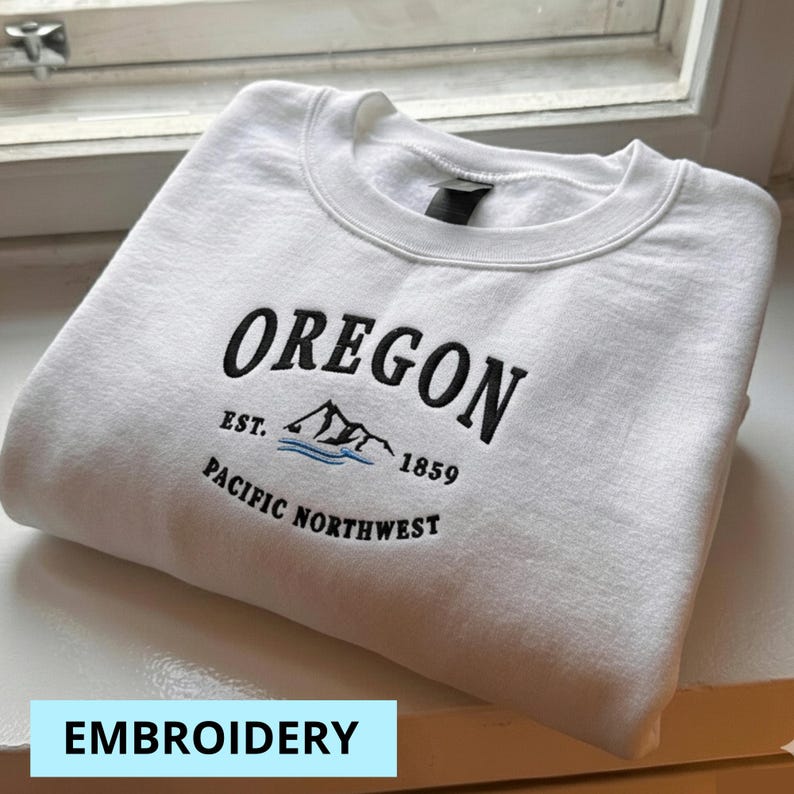 Pu&ograve; includere: Felpa bianca con la parola "OREGON" ricamata in nero, insieme a "EST. 1859" e "PACIFIC NORTHWEST". Il design include una grafica di montagna e acqua. La parola "EMBROIDERY" &egrave; in un rettangolo azzurro.
