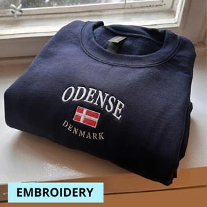 Puede incluir: Sudadera azul marino con texto bordado en blanco "ODENSE" sobre una bandera danesa y "DENMARK" debajo. La palabra "EMBROIDERY" está en un cuadro azul claro en la parte inferior de la imagen.