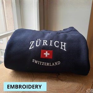 May include: A navy blue sweatshirt featuring white embroidered text: "ZÜRICH", a red square with a white cross, and "SWITZERLAND". The word "EMBROIDERY" is in a light blue rectangle at the bottom.