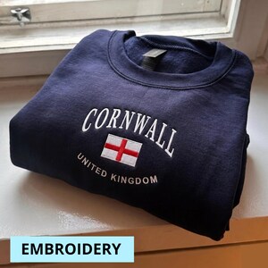 May include: Navy blue sweatshirt with white embroidered text reading "CORNWALL" above a flag and "UNITED KINGDOM" below. The flag has a white background with a red cross. The word "EMBROIDERY" is in a light blue box at the bottom.