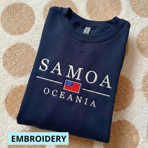 May include: Navy blue sweatshirt with white embroidered text reading "SAMOA OCEANIA" above a small flag of Samoa. The word "EMBROIDERY" is in a light blue rectangle at the bottom. The sweatshirt is folded on a patterned surface.