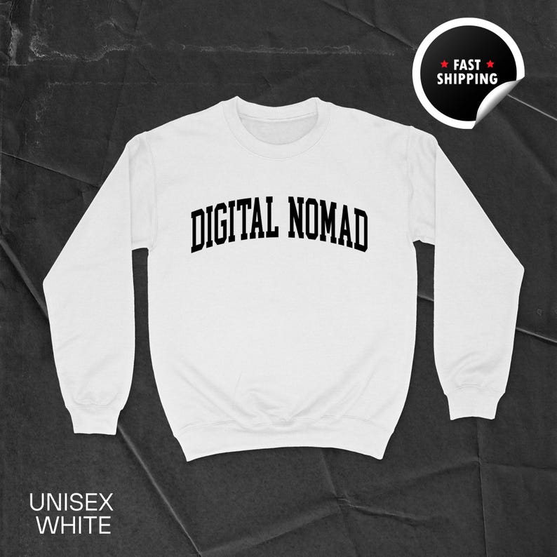 May include: White unisex sweatshirt with the text "DIGITAL NOMAD" in black, arched lettering. The sweatshirt has long sleeves and a crew neck. The words "UNISEX WHITE" are printed at the bottom. A black and white sticker with the words "FAST SHIPPING" is in the upper right corner.