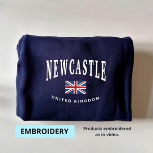 May include: A navy blue sweatshirt with white embroidery that reads "NEWCASTLE UNITED KINGDOM" and features a small embroidered Union Jack flag.