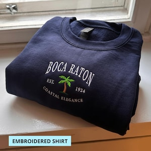 May include: Navy blue sweatshirt with white embroidered text that reads "Boca Raton" above a palm tree graphic and the text "Coastal Elegance". The text "Est. 1924" is also embroidered on the shirt. The words "Embroidered Shirt" are in a white box at the bottom.