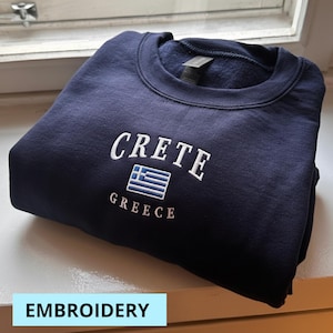 May include: Navy blue sweatshirt with white embroidered text reading "CRETE" above a Greek flag and "GREECE" below. The sweatshirt is folded neatly, showcasing the design. The word "EMBROIDERY" is in a light blue rectangle at the bottom.