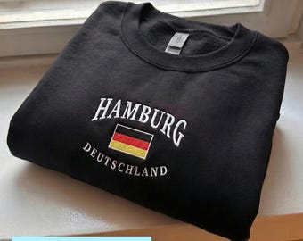 May include: Black crew neck sweatshirt with white embroidered text reading "HAMBURG" above a German flag and "DEUTSCHLAND" below. The word "EMBROIDERY" is in a light blue rectangle at the bottom.