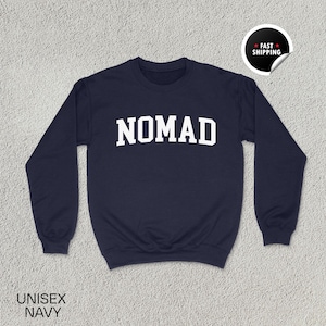 May include: Navy blue unisex sweatshirt with the word "NOMAD" in bold white letters across the chest. The sweatshirt has long sleeves and a crew neck. The words "UNISEX NAVY" are printed at the bottom left.