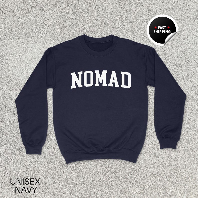 May include: Navy blue unisex sweatshirt with the word "NOMAD" in bold white letters across the chest. The sweatshirt has long sleeves and a crew neck. The words "UNISEX NAVY" are printed at the bottom left.