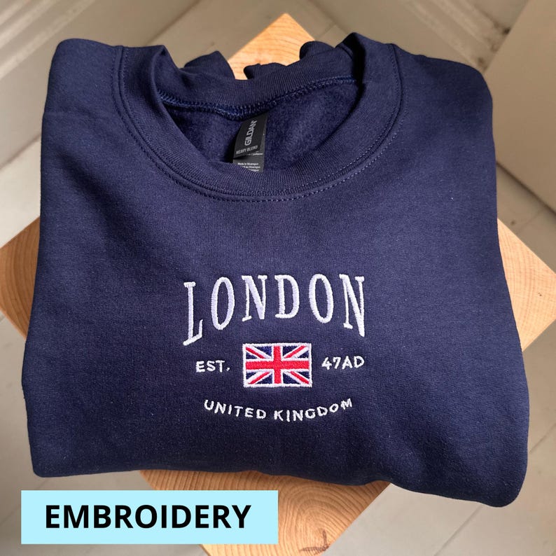 May include: Navy blue sweatshirt with white embroidered text that reads "LONDON EST. 47AD UNITED KINGDOM" and a red, white, and blue embroidered flag of the United Kingdom.