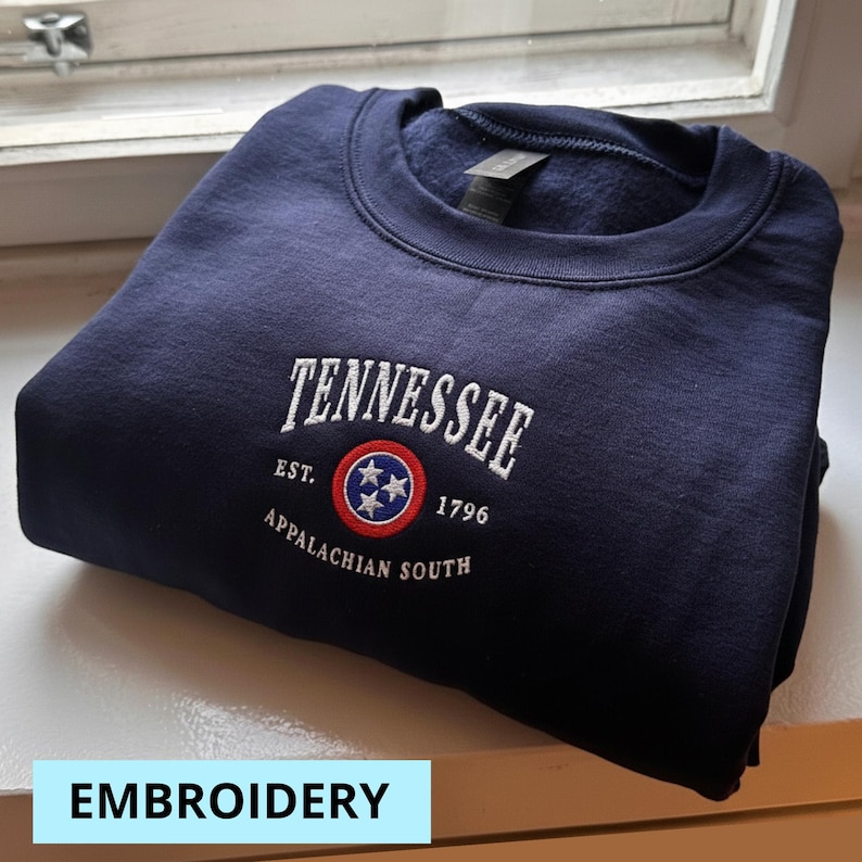 May include: Navy blue sweatshirt with white embroidered text reading "TENNESSEE" above a circular emblem and the text "EST. 1796 APPALACHIAN SOUTH". The sweatshirt is folded and resting on a windowsill. The word "EMBROIDERY" is in a light blue box.