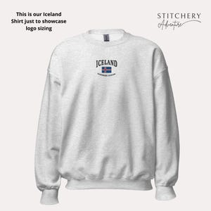Puede incluir: Una sudadera gris claro con la palabra "ICELAND" bordada en negro sobre una peque&ntilde;a bandera islandesa. El texto "northern europe" est&aacute; debajo de la bandera. La sudadera tiene mangas largas y cuello redondo.