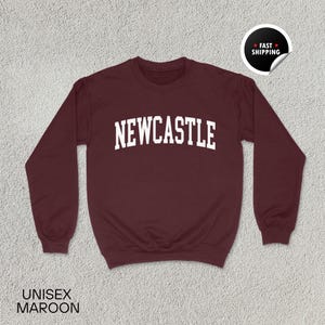 Puede incluir: Sudadera burdeos con la palabra "NEWCASTLE" en letras blancas. La sudadera tiene mangas largas y cuello redondo. Las palabras "UNISEX MAROON" est&aacute;n impresas en la parte inferior. Una pegatina de "FAST SHIPPING" est&aacute; en la esquina superior derecha.