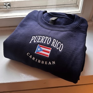 May include: A navy blue crewneck sweatshirt featuring white embroidered text that reads Puerto Rico above a small, rectangular Puerto Rican flag graphic, with the word Caribbean embroidered in white text below the flag on the chest.