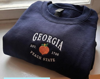 Könnte beinhalten: Ein marineblaues Sweatshirt mit dem weiß gestickten Wort "GEORGIA", einer Pfirsichgrafik und dem Text "EST. 1788 PEACH STATE". Das Sweatshirt ist gefaltet und auf einer weißen Oberfläche präsentiert.
