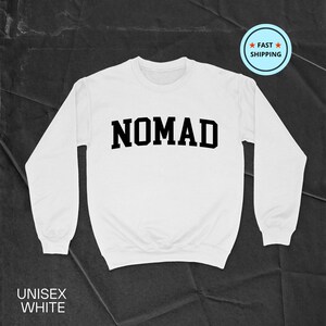 May include: White unisex sweatshirt with the word "NOMAD" in bold, black letters across the chest. The sweatshirt has long sleeves and a crew neck. The words "UNISEX WHITE" are printed at the bottom. A "FAST SHIPPING" badge is in the upper right corner.