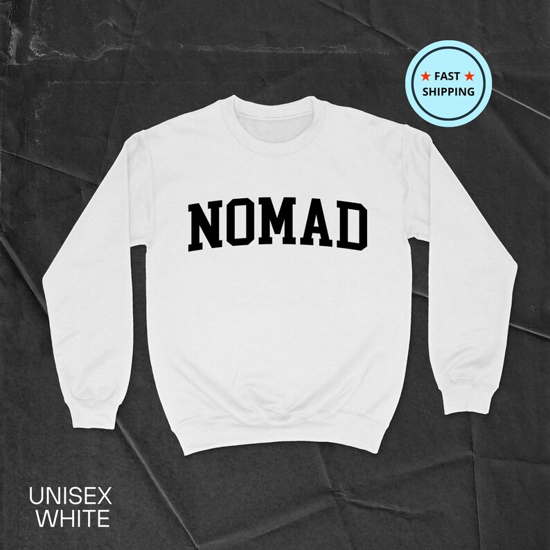 May include: White unisex sweatshirt with the word "NOMAD" in bold, black letters across the chest. The sweatshirt has long sleeves and a crew neck. The words "UNISEX WHITE" are printed at the bottom. A "FAST SHIPPING" badge is in the upper right corner.