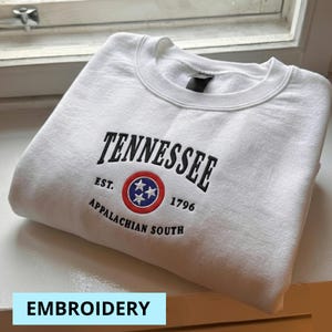 May include: White crewneck sweatshirt with the word "Tennessee" embroidered in black arched above a circular emblem with three white stars on a blue background, surrounded by a red ring. The text "EST. 1796 APPALACHIAN SOUTH" is below the emblem.