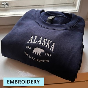 May include: A navy blue sweatshirt with white embroidered text and a bear graphic. The word "ALASKA" is arched above "EST. 1959" and "THE LAST FRONTIER". The word "EMBROIDERY" is in a light blue rectangle at the bottom.