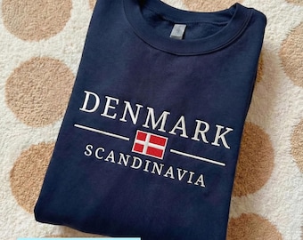 Op de afbeelding: Een marineblauw sweatshirt met het woord "DENMARK" in het wit geborduurd, samen met de Deense vlag en het woord "SCANDINAVIA". Het woord "EMBROIDERY" staat onderaan de afbeelding.