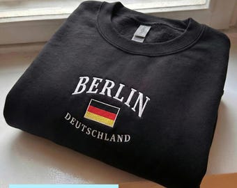 May include: Black crewneck sweatshirt with white embroidered text "BERLIN" above a small German flag and the word "DEUTSCHLAND". The sweatshirt is folded on a white surface. The word "EMBROIDERY" is in a light blue box at the bottom.