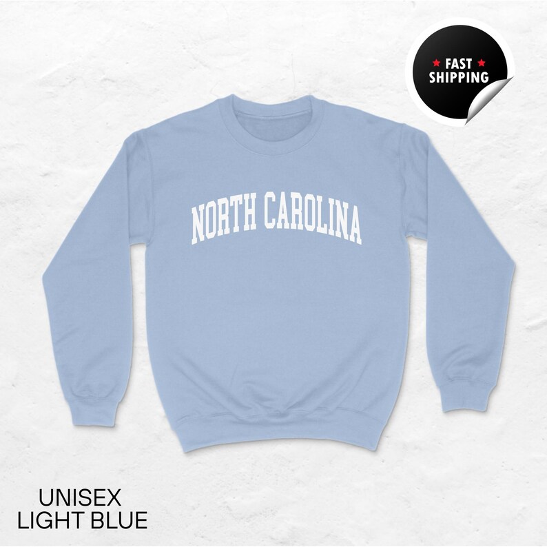 May include: Light blue unisex sweatshirt with the text "NORTH CAROLINA" in white, arched across the chest. The sweatshirt is a solid light blue color. The words "UNISEX LIGHT BLUE" are printed at the bottom.