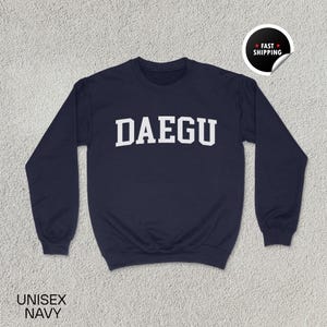 May include: Navy blue unisex sweatshirt with the word "DAEGU" in white block letters. The sweatshirt has long sleeves and a crew neck. The words "UNISEX NAVY" are printed at the bottom left of the image. A "FAST SHIPPING" sticker is in the top right corner.