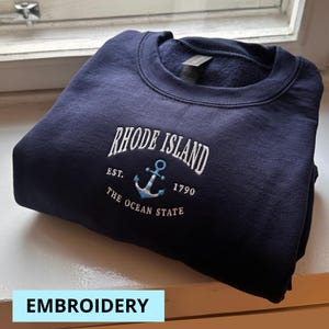 Può includere: Felpa blu navy con ricamo bianco "RHODE ISLAND" sopra un'ancora e la scritta "EST. 1790 THE OCEAN STATE". La felpa è piegata e appoggiata su una superficie bianca.