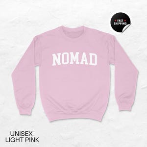 May include: A light pink unisex sweatshirt with the word "NOMAD" in white block letters across the chest. The sweatshirt has long sleeves and a crew neck. The words "UNISEX LIGHT PINK" are printed at the bottom.