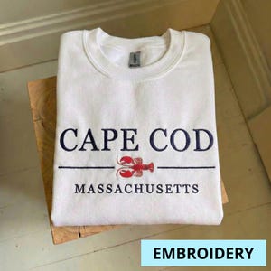 Puede incluir: Sudadera blanca de cuello redondo con el texto bordado "CAPE COD MASSACHUSETTS" en azul marino. Un gráfico de langosta roja está centrado debajo del texto. La palabra "EMBROIDERY" está en un cuadro azul claro en la parte inferior.