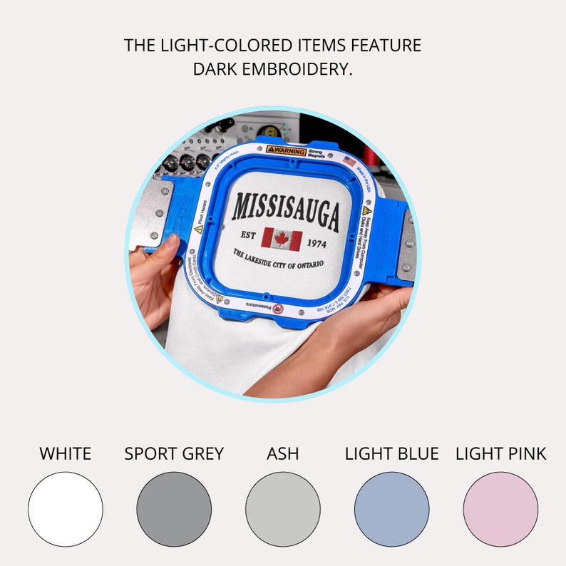 May include: A blue embroidery hoop holds a white fabric with the text "MISSISSAUGA EST 1974 THE LAKESIDE CITY OF ONTARIO" in black. The image also shows color swatches: white, sport grey, ash, light blue, and light pink.