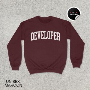May include: Maroon unisex sweatshirt with the word "DEVELOPER" in white, bold, block letters across the chest. The sweatshirt has a crew neck and long sleeves. A "Fast Shipping" sticker is in the upper right corner.