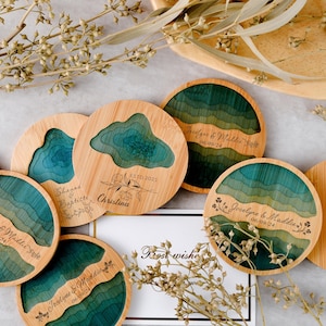May include: A collection of round wooden coasters with a layered design in shades of teal and green. Some coasters feature engraved names and dates, while others have a cutout design. The coasters are arranged with dried floral accents.