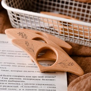 May include: Wooden thumb page holders with floral designs. These book accessories are light brown and feature a circular finger hole. The page holders are resting on a book with text and a brown cloth.