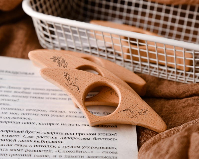 May include: Wooden thumb page holders with floral designs. These book accessories are light brown and feature a circular finger hole. The page holders are resting on a book with text and a brown cloth.