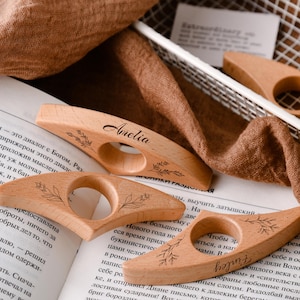 May include: Wooden thumb page holders with engraved names and floral designs. The light-colored wood features a circular opening for the thumb and is resting on the pages of an open book. The names "Amelia" and "Finley" are visible.