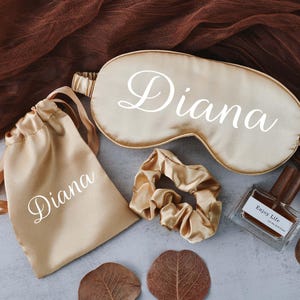 May include: A set of personalised gifts in a gold satin colour. The set includes a sleep mask, a small drawstring bag, a hair scrunchie, and a bottle of perfume. All items are personalised with the name "Diana".